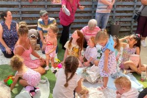Families of Ash Lodge Nursing Home gathered on a large picnic rug in the garden. Children are anticipating a game of pass the parcel.